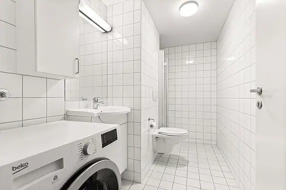 Flislagt baderom med dusj, veggmontert toalett, servantskap og opplegg for vaskemaskin. Varmekabler i gulv